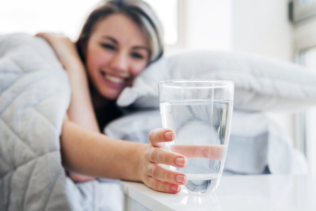Lächelnde Frau im Bett greift nach einem Glas frischem Wasser auf dem Nachttisch, um hydriert in den Tag zu starten.