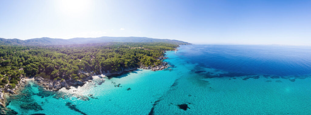 Eine sonnige Panorama-Luftaufnahme der griechischen Küste mit üppig grünen Wäldern und kristallklarem, türkisfarbenem Wasser.