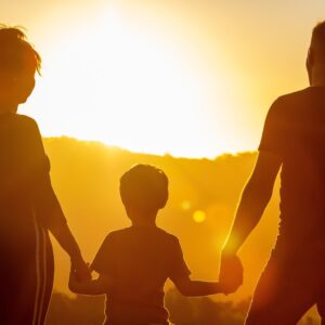 Eine Familie mit Vater, Mutter und Kind hält sich bei Sonnenuntergang an den Händen und blickt auf eine leuchtende Berglandschaft.