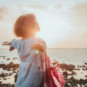 Eine Frau streckt bei Sonnenuntergang am felsigen Meerufer die Arme aus und geniesst einen Moment der Freiheit und Lebensfreude.
