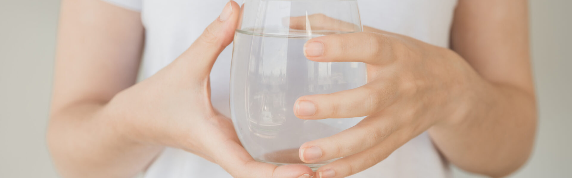 Eine Frau in einem weißen T-Shirt hält ein Glas mit klarem, reinem Wasser in beiden Händen.