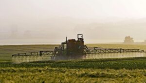 Ein Traktor mit breiten Sprüharmen fährt durch ein grünes Getreidefeld und versprüht großflächig Pestizide in der diesigen Morgenluft.
