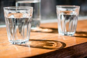 Drei Gläser mit klarem Wasser auf einem Holztisch, das vorderste Glas im Fokus.