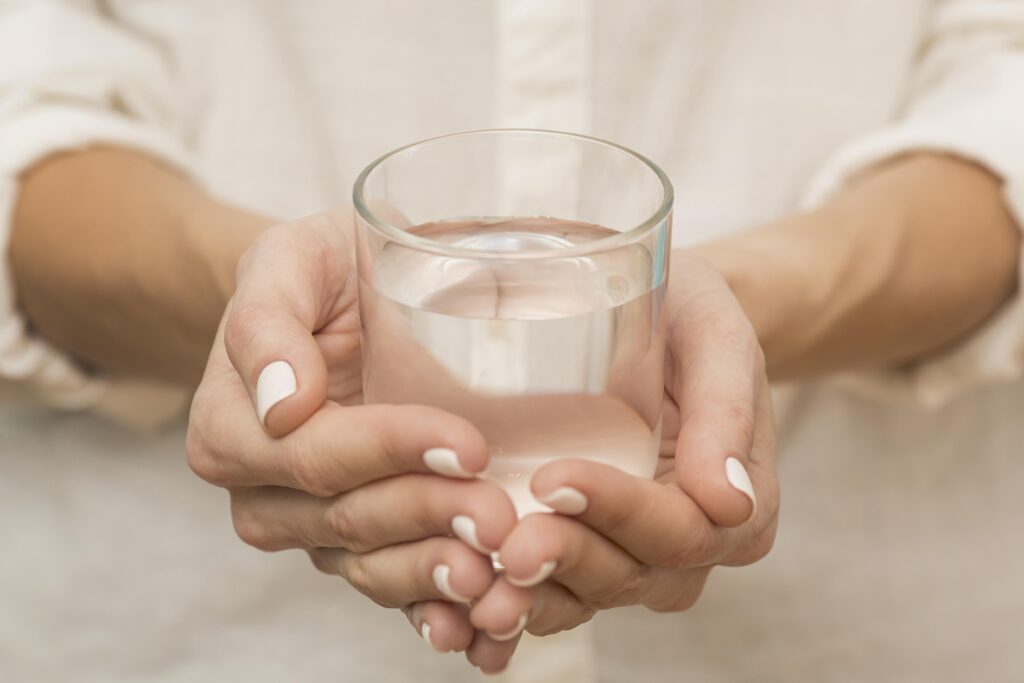 Eine Frau in einem weißen Hemd hält mit beiden Händen ein Glas mit klarem, reinem Wasser.