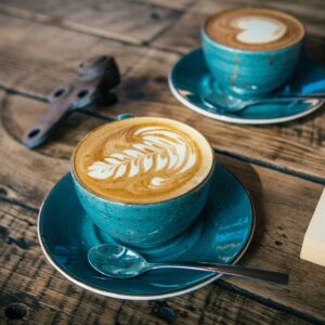 Zwei türkisfarbene Tassen Cappuccino mit Latte Art stehen auf einem rustikalen Holztisch neben einem aufgeschlagenen Buch und schaffen eine gemütliche Atmosphäre.